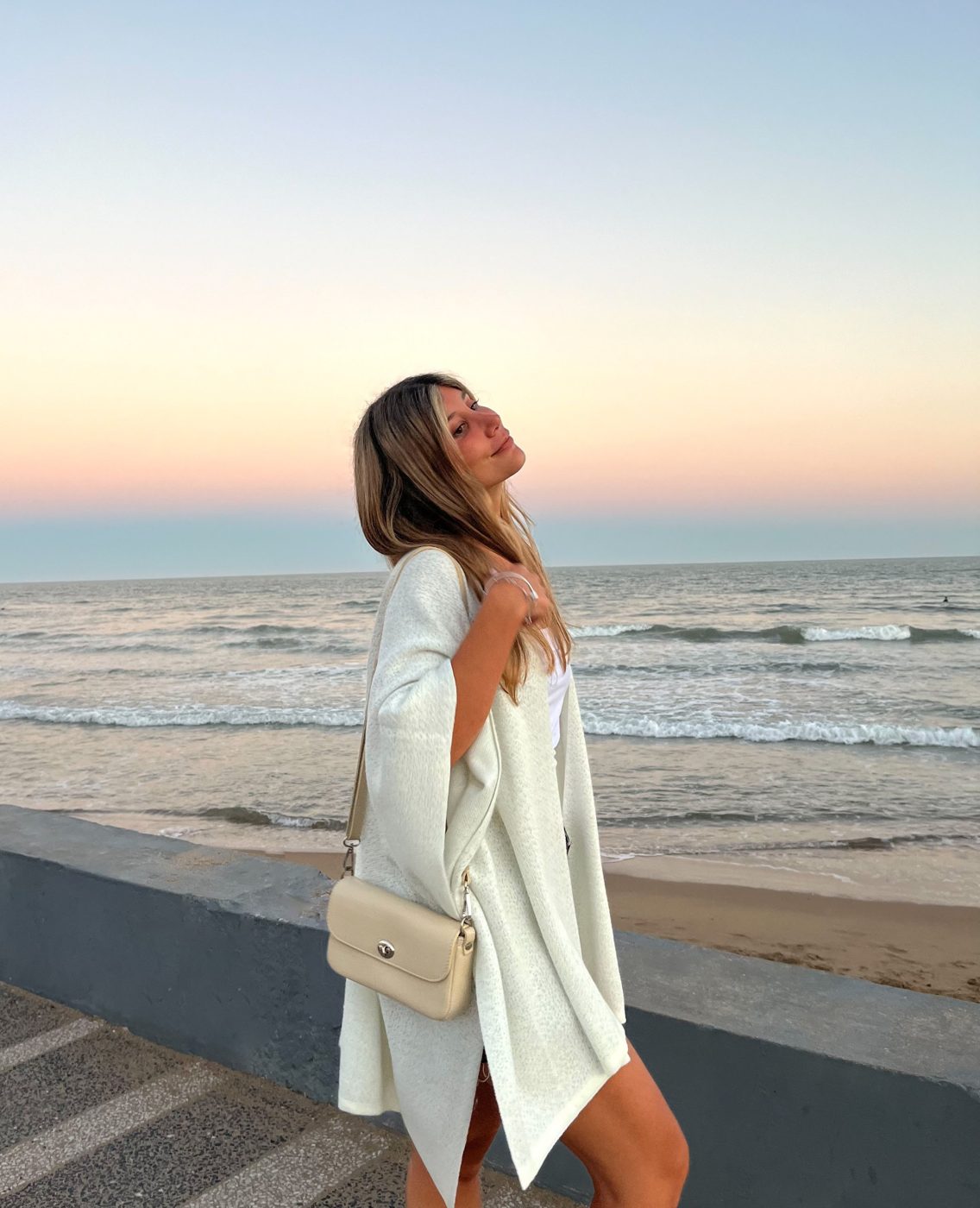 Victoria en la rambla al atardecer, con saco blanco y cartera beige, frente al mar
